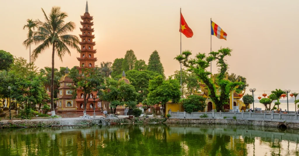 Showing the historic Trấn Quốc Pagoda in Hanoi, Vietnam, featuring a tall, multi-tiered red stupa surrounded by lush trees, flags, and a calm body of water during a warm, hazy sunset or sunrise | KASS Malaysia