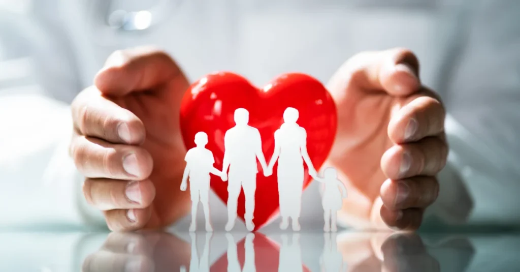 A pair of adult hands surrounds a bright red heart, while in front of the heart is a white silhouette of a family of four. Two adults and two children holding hands.