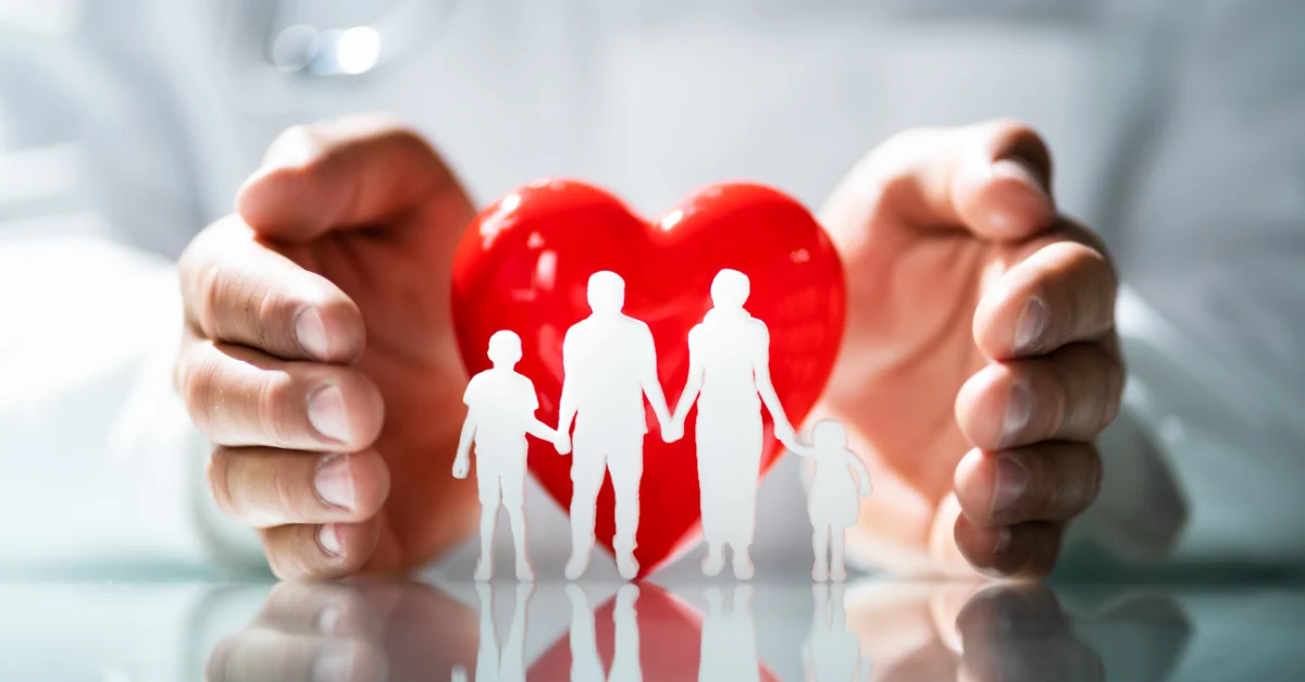 A pair of adult hands surrounds a bright red heart, while in front of the heart is a white silhouette of a family of four. Two adults and two children holding hands.